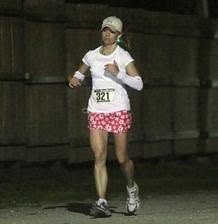 Night photo from my first 24-hour run: bib 321, white long sleeves, red flowered skirt, mid-stride on the track.
