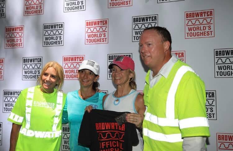 Four of us posing in front of a red-and-white 'Badwater World's Toughest' backdrop, two crew in hi-vis yellow, holding up a black Badwater official finisher shirt.