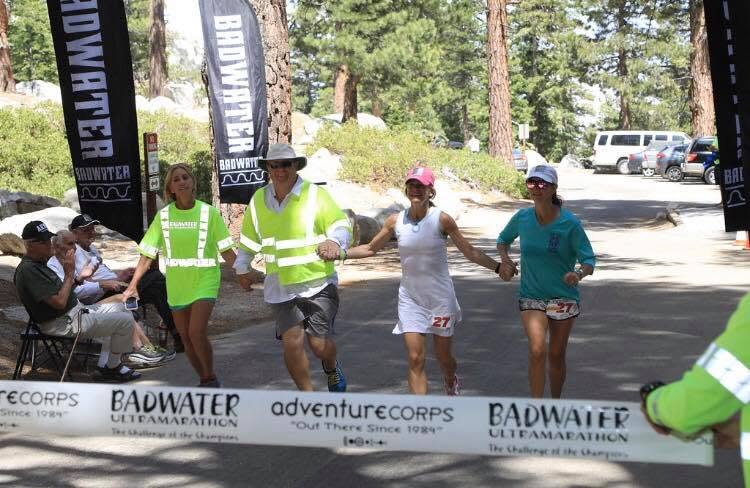 Crossing the finish line in a pine forest, holding hands with two other runners under the Badwater Ultramarathon and adventureCORPS tape; I'm in white with bib number 27 and a pink cap.