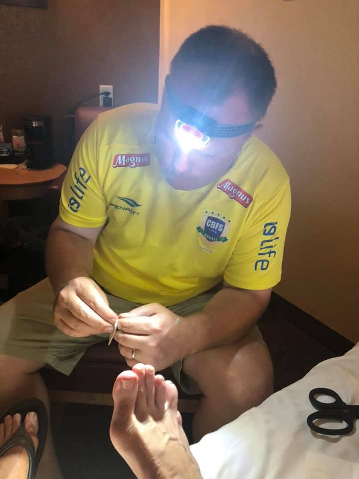 A crew member in a yellow jersey and headlamp hunched over in a hotel room, carefully working on my bare foot, with scissors resting on the bed beside us.
