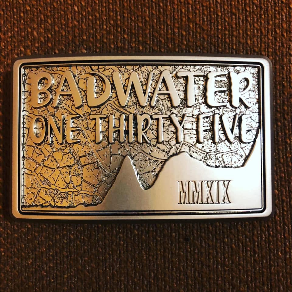 Close-up of the silver finisher's belt buckle reading 'BADWATER ONE THIRTY FIVE MMXIX' over a cracked-earth desert texture and a mountain silhouette.