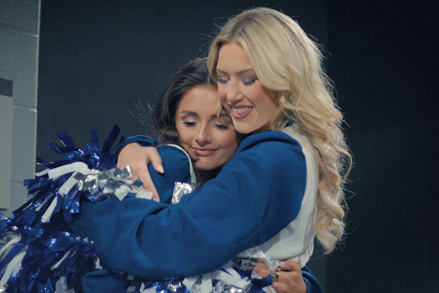 Two Dallas Cowboys Cheerleaders in navy and white uniforms share a warm hug, eyes closed and smiling, holding blue and silver pom-poms against a dark backstage backdrop.