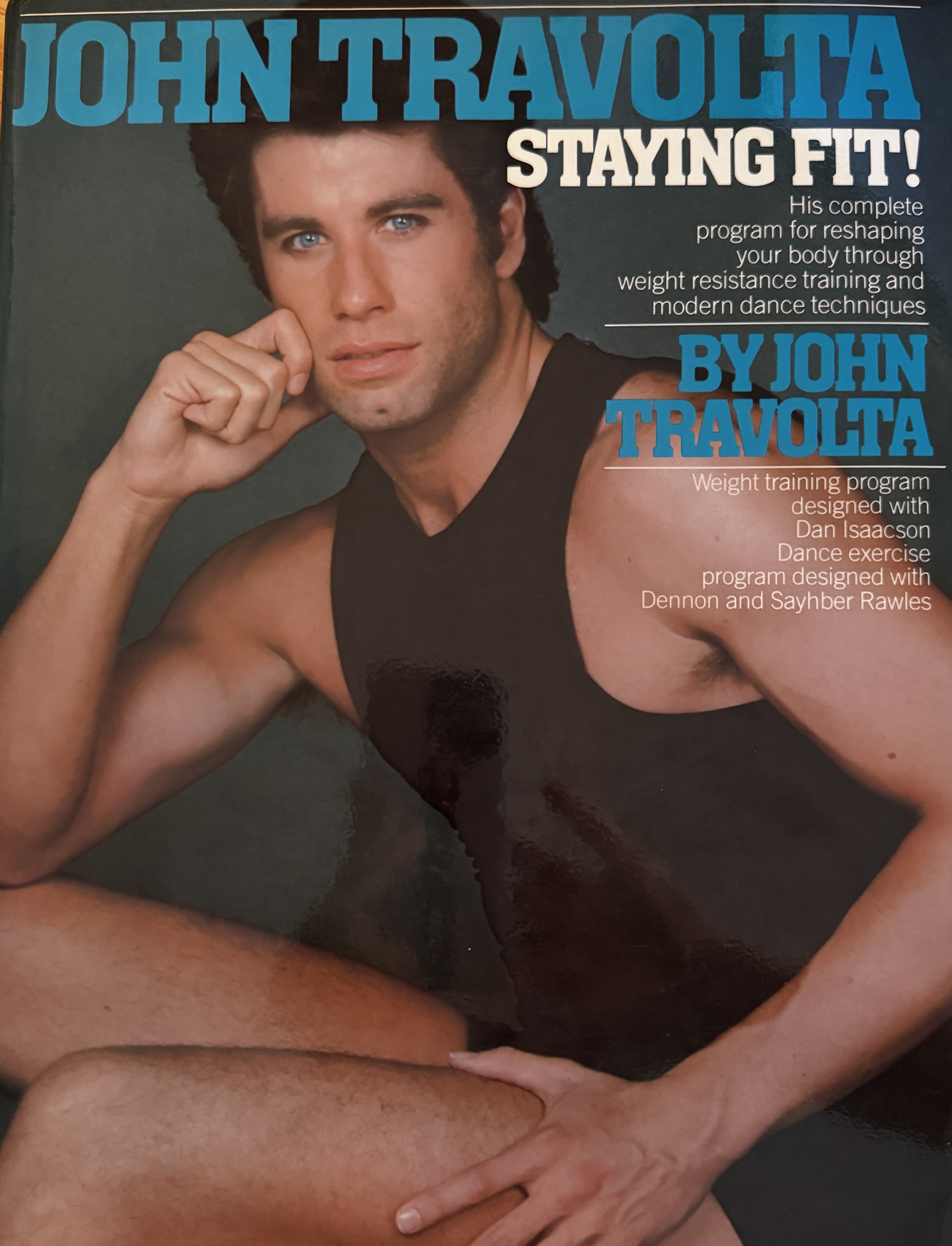 Cover of the 1984 book 'John Travolta: Staying Fit!' A young John Travolta sits in a black sleeveless leotard, chin resting on one hand, gazing at the camera. Blue and cream cover text reads 'Staying Fit! His complete program for reshaping your body through weight resistance training and modern dance techniques.'