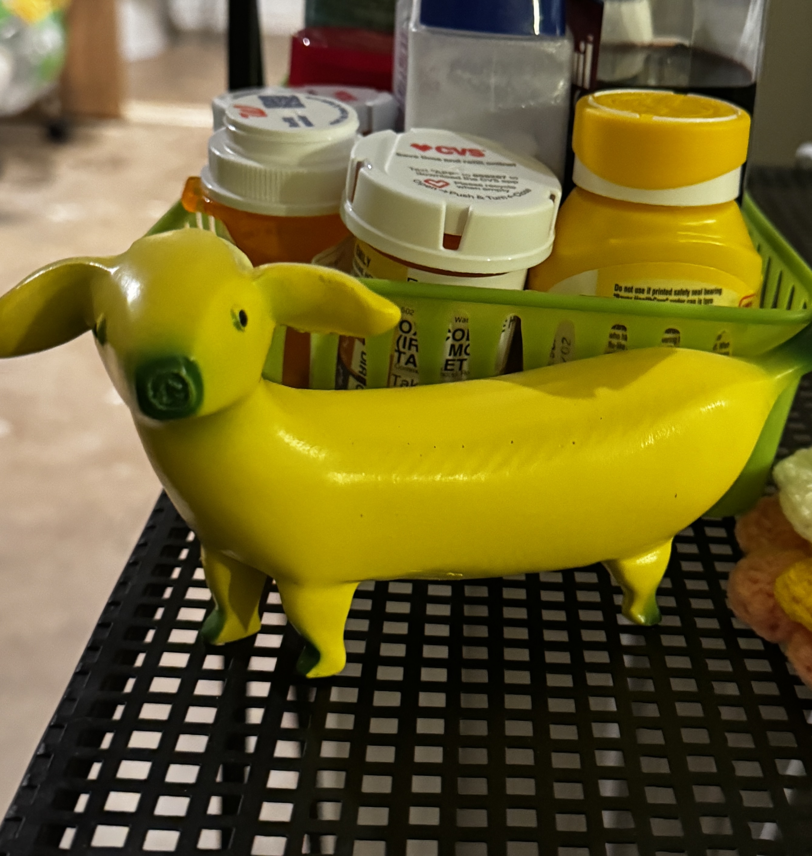 A bright yellow figurine of a dachshund whose body is a banana, with little ears and a goofy snout, standing on a black grid surface in front of a green basket stuffed with prescription pill bottles and a honey-colored bottle.