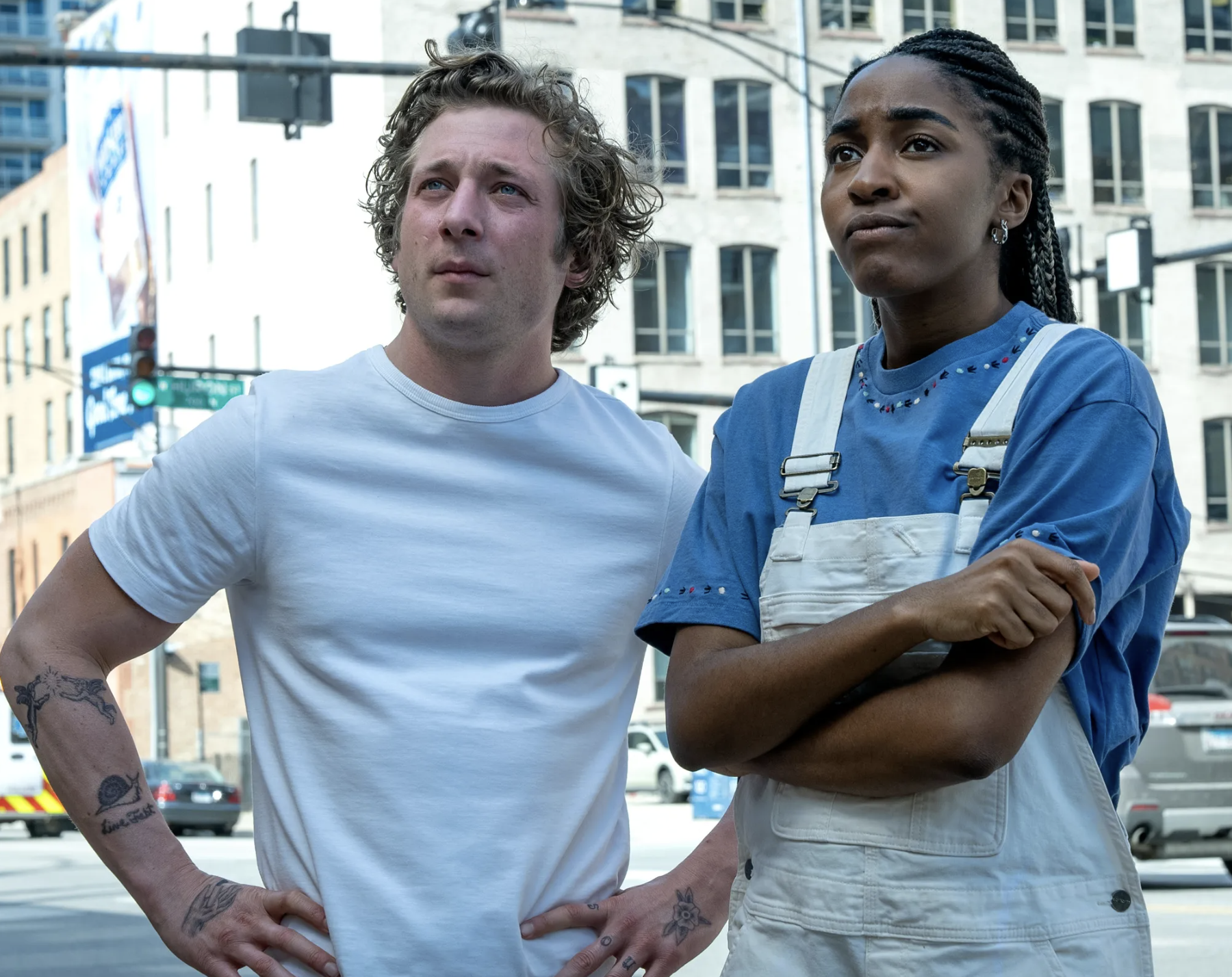 Two characters from The Bear stand on a Chicago street. On the left, a curly-haired man in a white t-shirt with forearm tattoos rests his hands on his hips. On the right, a woman in a blue shirt and white overalls stands with her arms crossed, both looking off to the side.