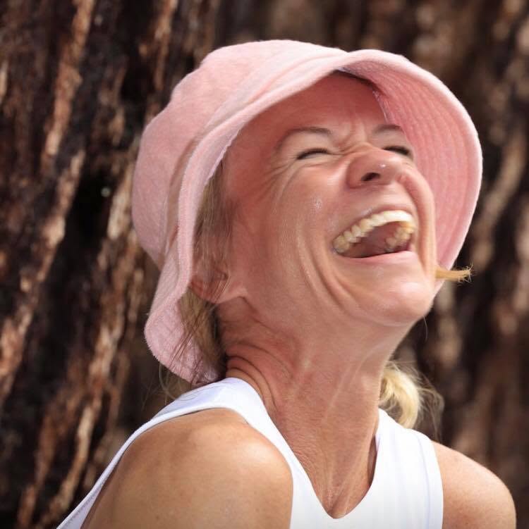 A woman in a pink bucket hat and white tank top, head thrown back mid-laugh against a tree trunk.