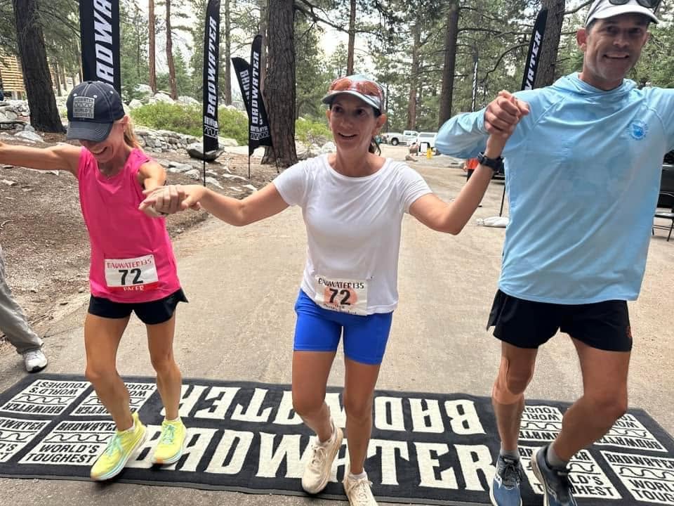 Three people crossing the Badwater finish mat at Whitney Portal, hands raised and clasped together in celebration among the pines and race flags.