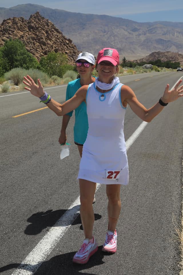 A grinning runner in a white dress with bib 27, arms flung wide on a desert road below the Alabama Hills, a second runner in teal just behind.
