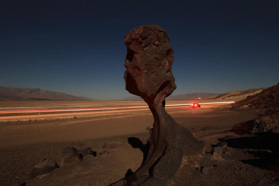 Night long-exposure of Mushroom Rock in Death Valley, red and white car light-trails streaking along the road behind it under a star-scattered sky.