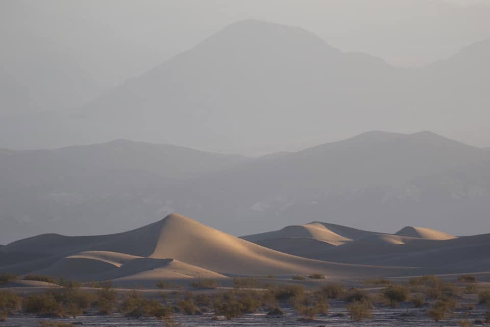 Mesquite Flat sand dunes at first light, soft golden crests layered against hazy blue-gray mountain ridges.
