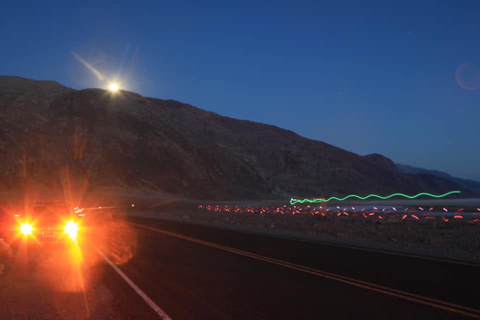 A dark desert road at night: a car's headlights flare at left while squiggles of red and green runner safety-lights trail off into the distance below a moonlit ridge.