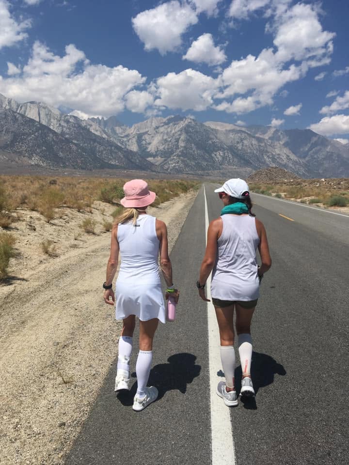 Two runners in white dresses and tall compression socks walking away down an empty desert highway toward the jagged Sierra Nevada.