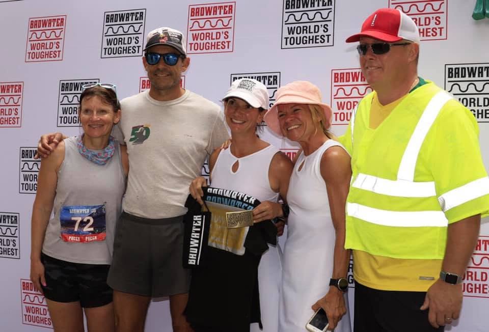 Five people posed at the 'Badwater — World's Toughest' backdrop; the runner in the center holds a finisher shirt and belt buckle, a volunteer in a hi-vis vest at the end.
