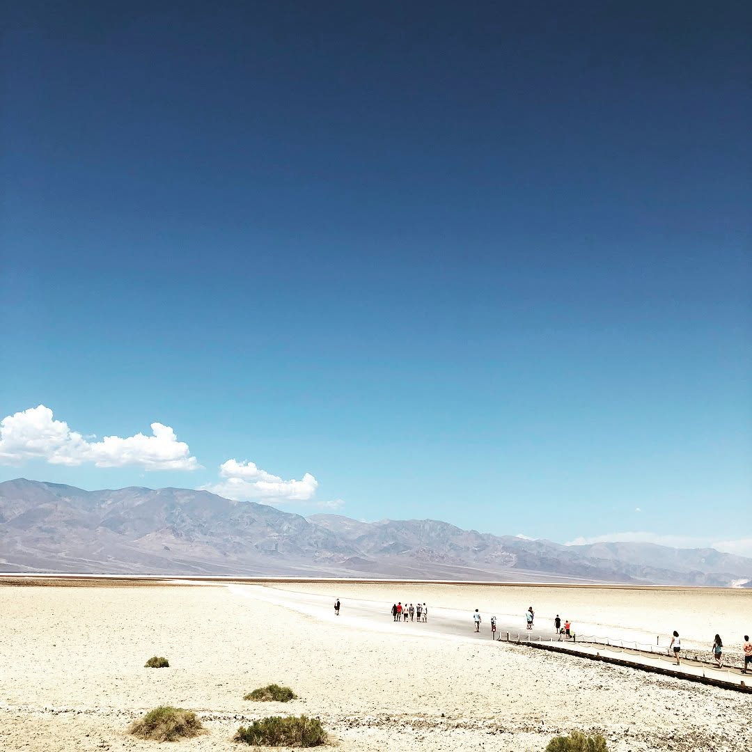 The vast white salt flats of Badwater Basin under a deep blue sky, tiny figures strung along the boardwalk with brown mountains behind.