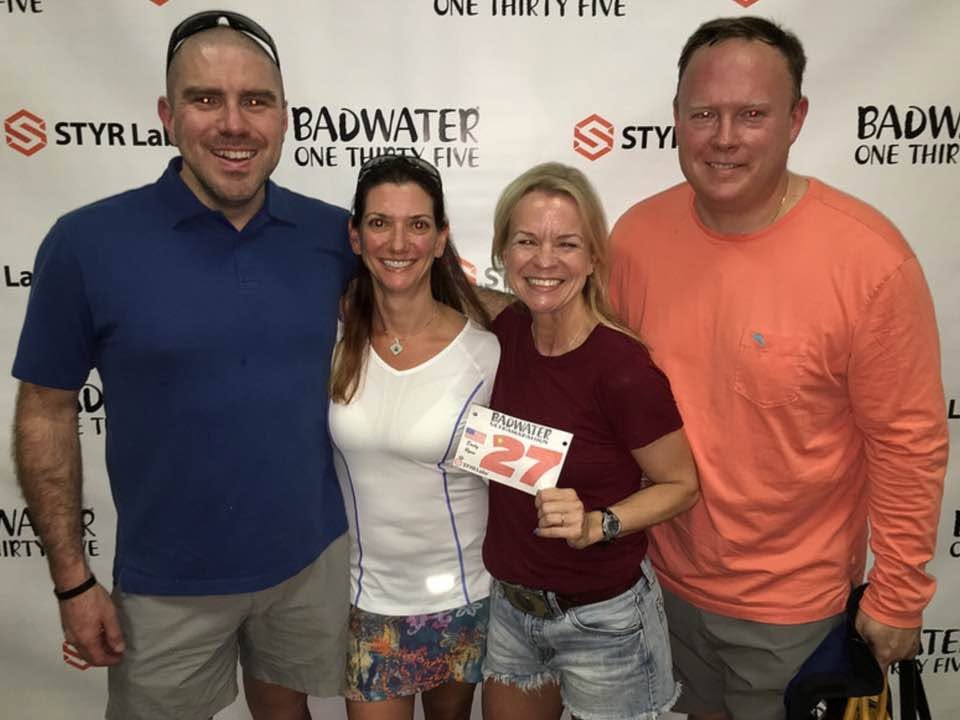 Four people at the 'Badwater One Thirty Five' backdrop; a woman in a maroon shirt holds up race bib number 27.