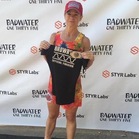A runner in a red cap and tie-dye dress holding up a black 'Badwater Official Finisher' shirt and her buckle at the backdrop.