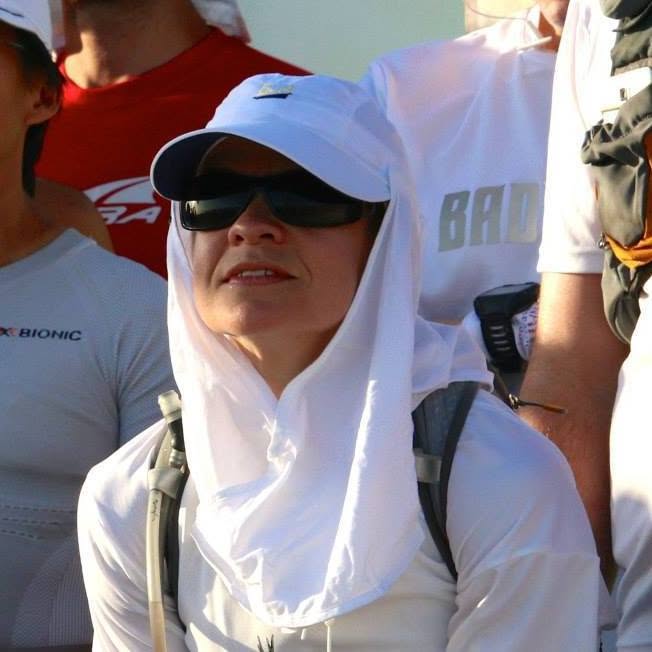 Close-up of a runner in sunglasses and a white cap with a neck sun-flap, hydration pack on, packed in among other runners in white sun gear under hard light.