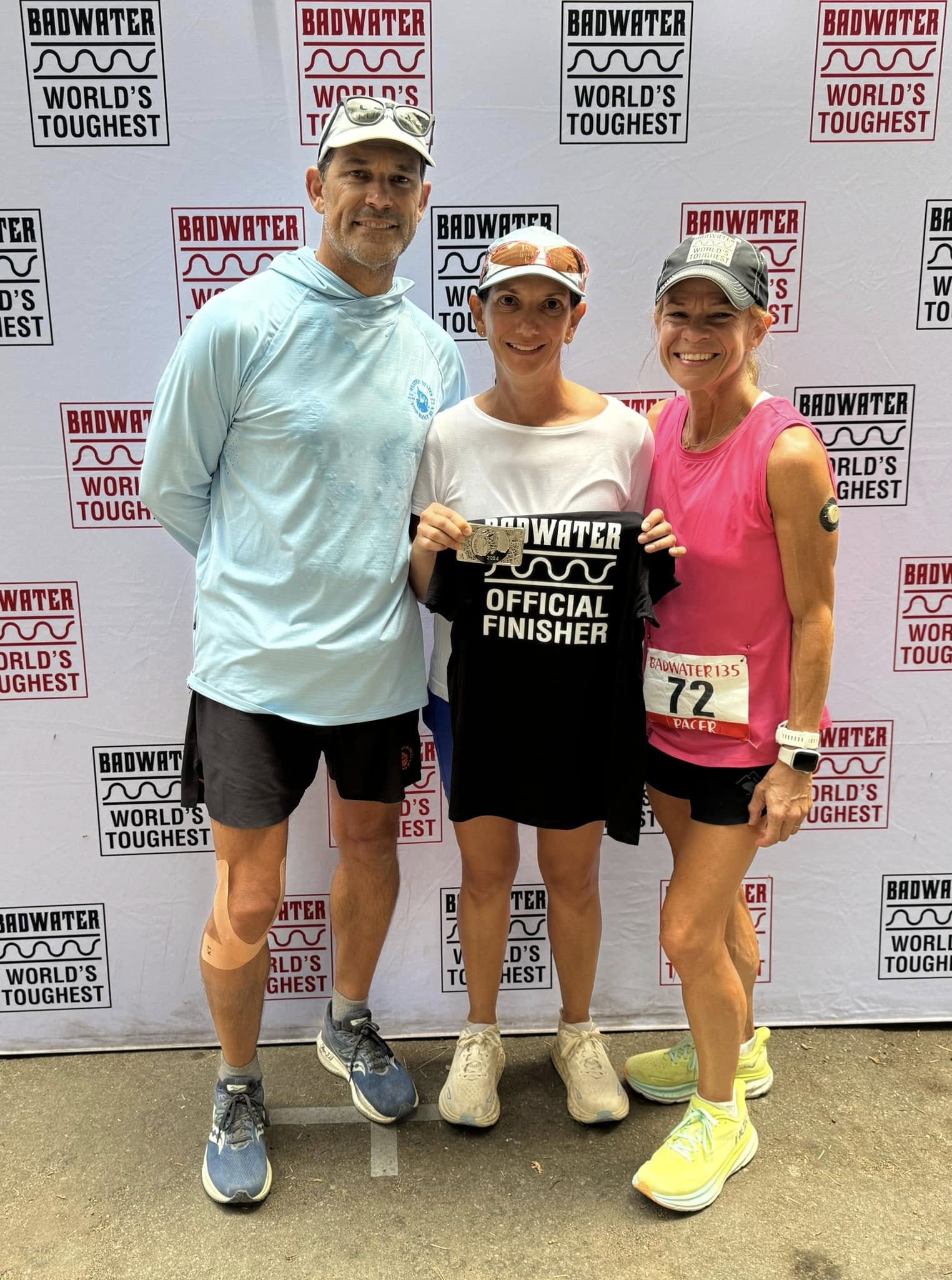 Three at the 'Badwater — World's Toughest' backdrop; the finisher in the center holds up her shirt and buckle, a pacer wearing bib 72 beside her.