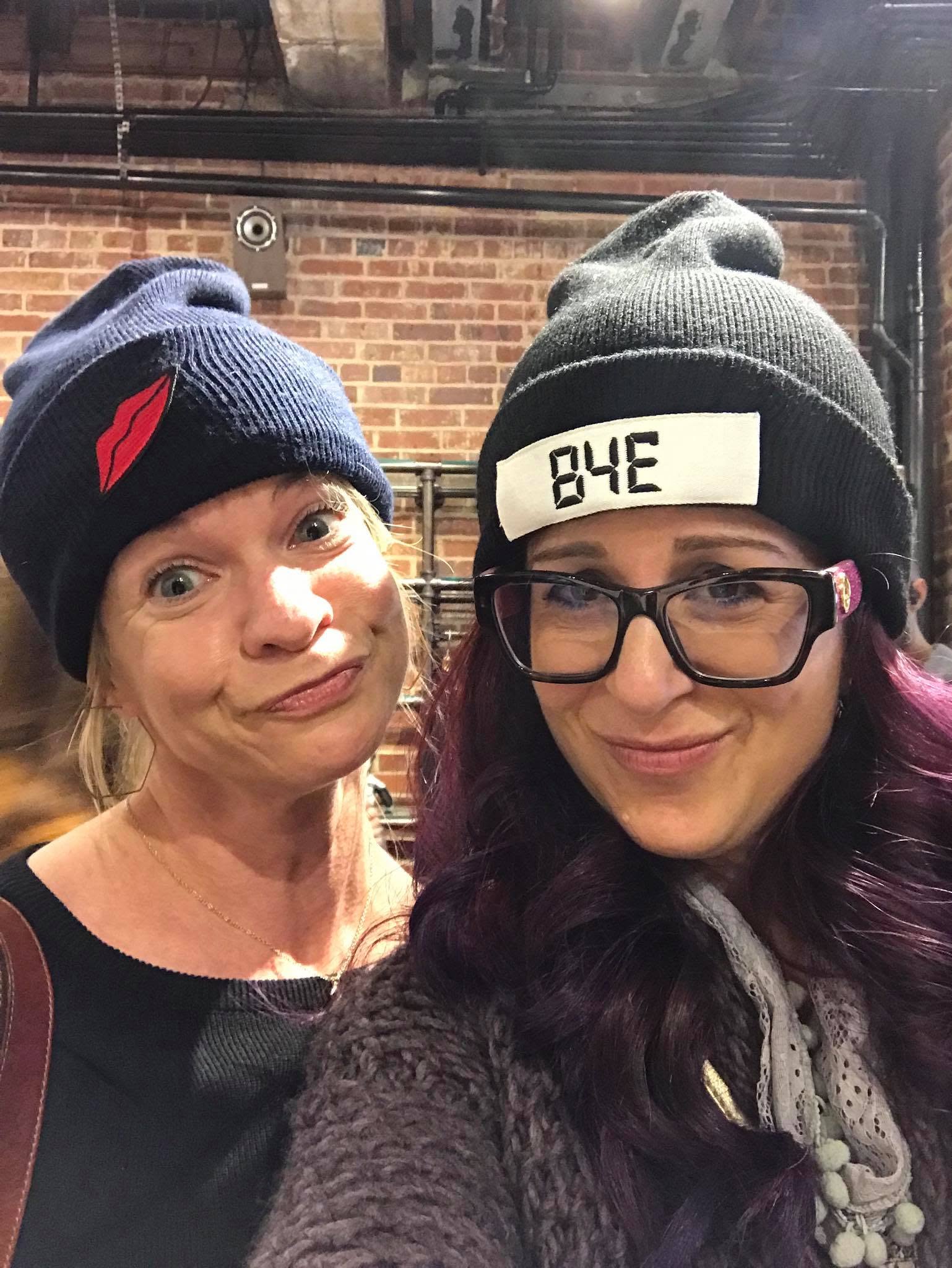 Two smiling women posing cheek to cheek in knit beanies against an exposed-brick and pipe wall; one beanie has a red lips patch, the other reads 'BYE' in a digital-clock font.