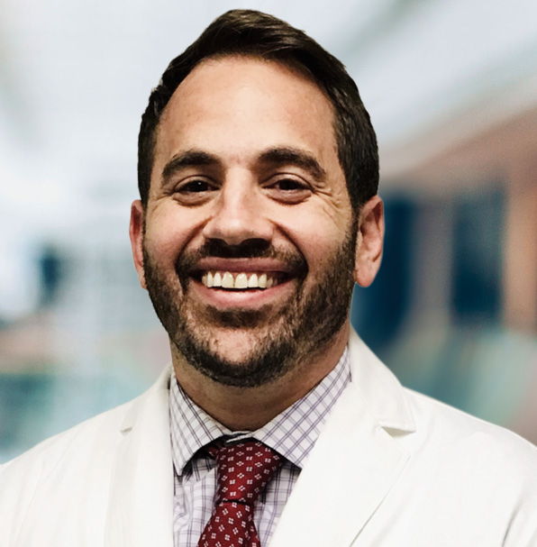 Dr. Ben Stein: a smiling man with short dark hair and a trimmed dark beard, wearing a white physician's coat over a checked dress shirt and a dark red patterned tie, photographed in a softly blurred hospital corridor.