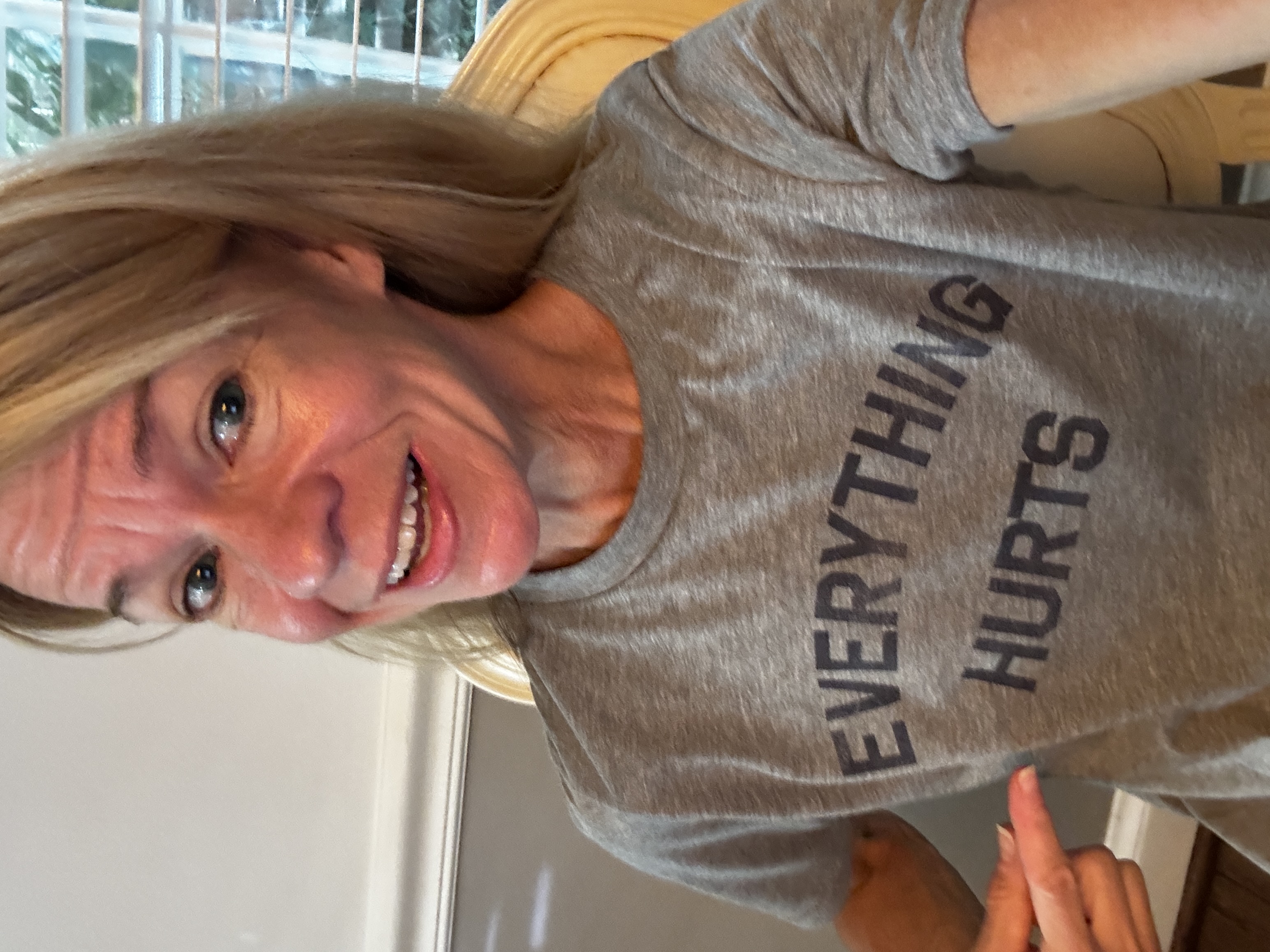 Selfie of a smiling woman with long blond hair, wearing a heather-gray t-shirt printed with bold black block letters reading 'EVERYTHING HURTS.'