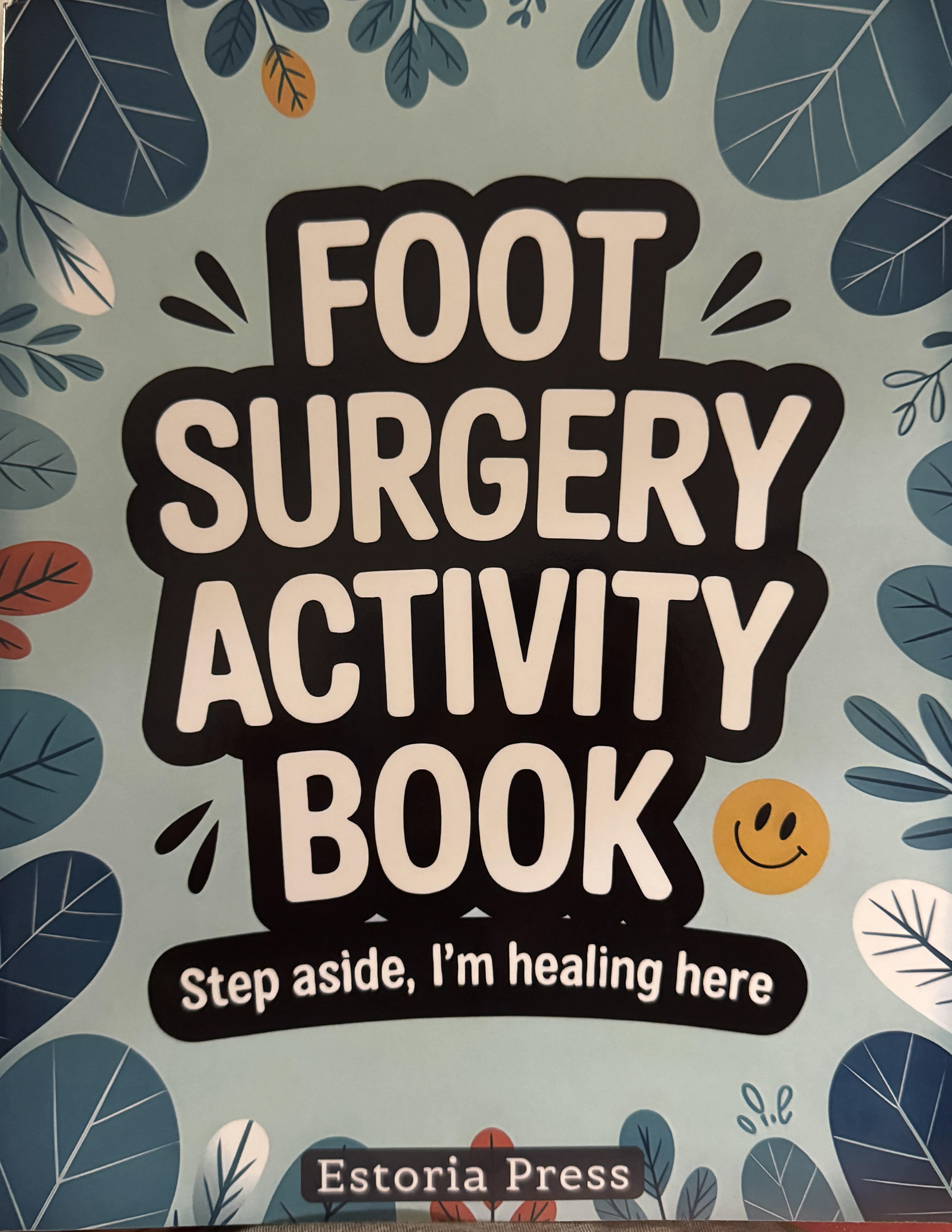 Cover of the 'Foot Surgery Activity Book' by Estoria Press. Bold white block letters on a black sticker-style outline spell the title against a soft blue background scattered with illustrated eucalyptus leaves. A small smiley face sits beside the word BOOK, and a black banner underneath reads 'Step aside, I'm healing here.'