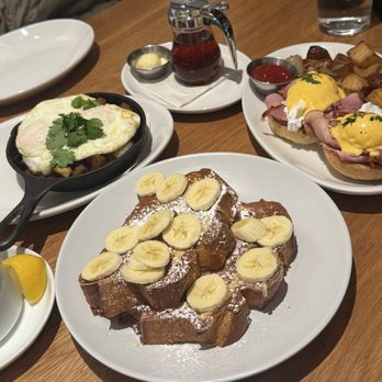 A restaurant table set with brunch: a plate of thick French toast topped with sliced bananas and powdered sugar in front, a skillet hash with a fried egg, and eggs Benedict with a side of potatoes in the background, plus a small pitcher of maple syrup.
