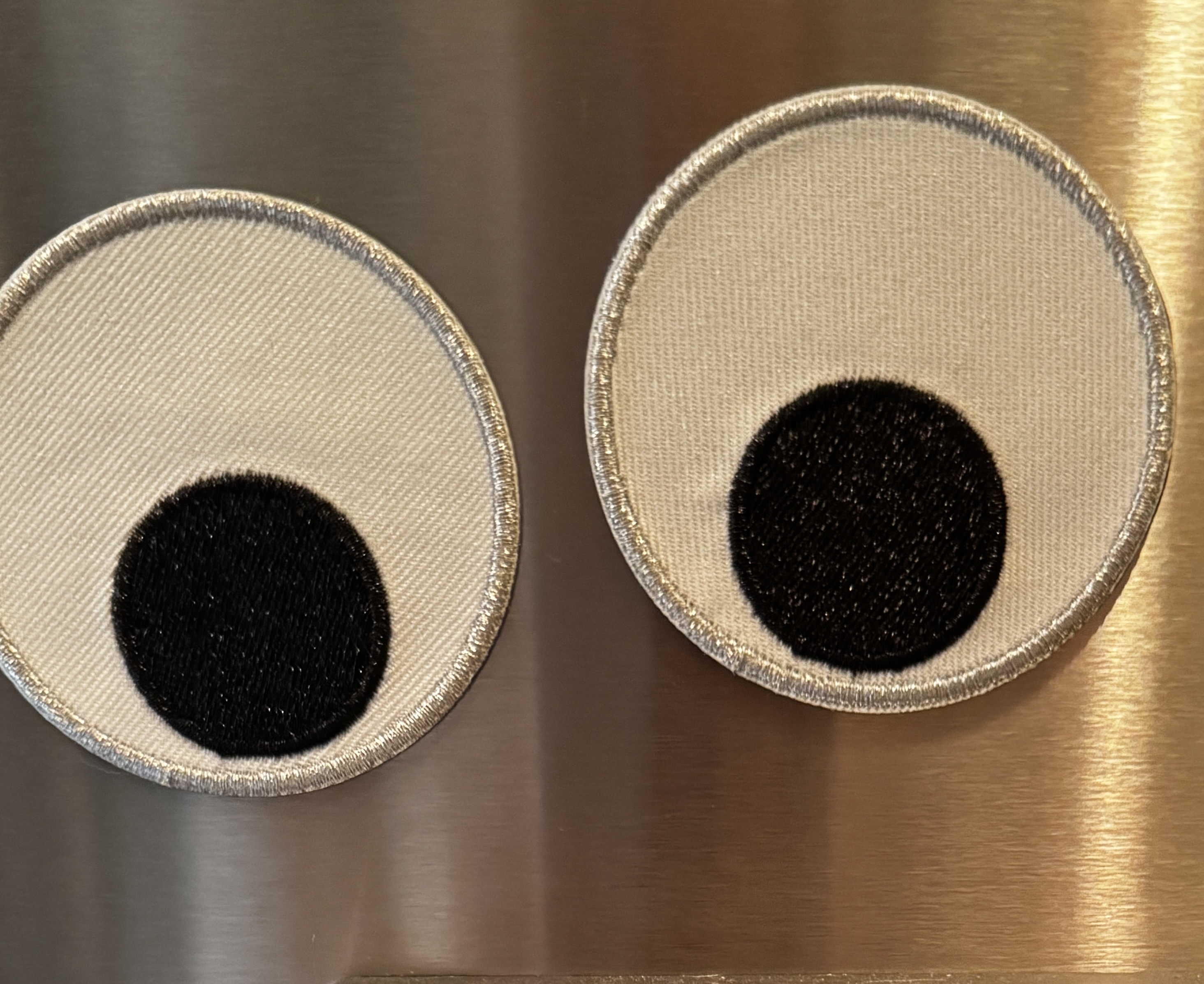 Two oversized embroidered googly-eye magnets stuck on a stainless steel refrigerator: round cream-white fabric patches with silver borders and big black pupils, both glancing downward.