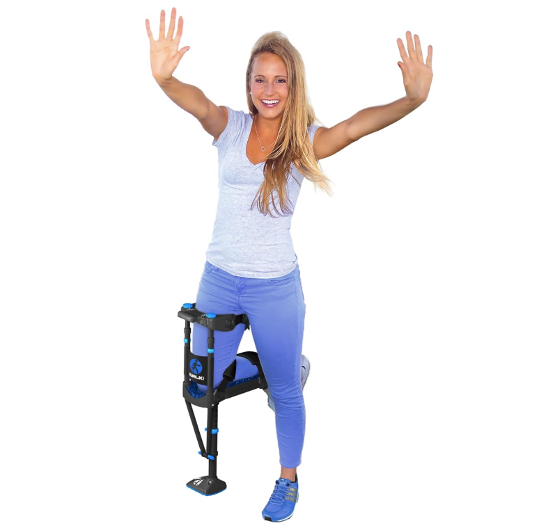 A smiling woman in a gray t-shirt and blue jeans throws both arms up in the air, hands free, with a black-and-blue iWALK hands-free crutch strapped to her bent left leg and a blue sneaker on her standing right foot.