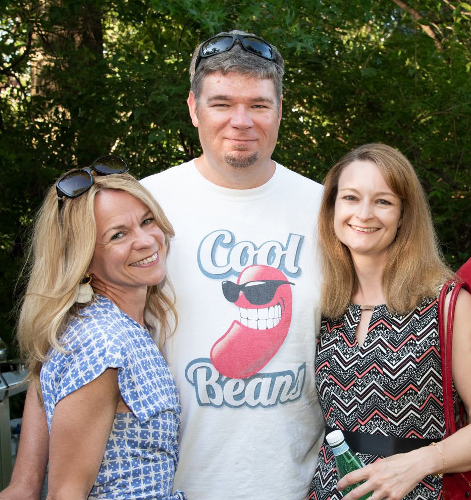 Three friends posed together outdoors in front of green summer trees; the man in the center wears a white 'Cool Beans' t-shirt with a sunglasses-and-tongue graphic, a woman on either side of him.
