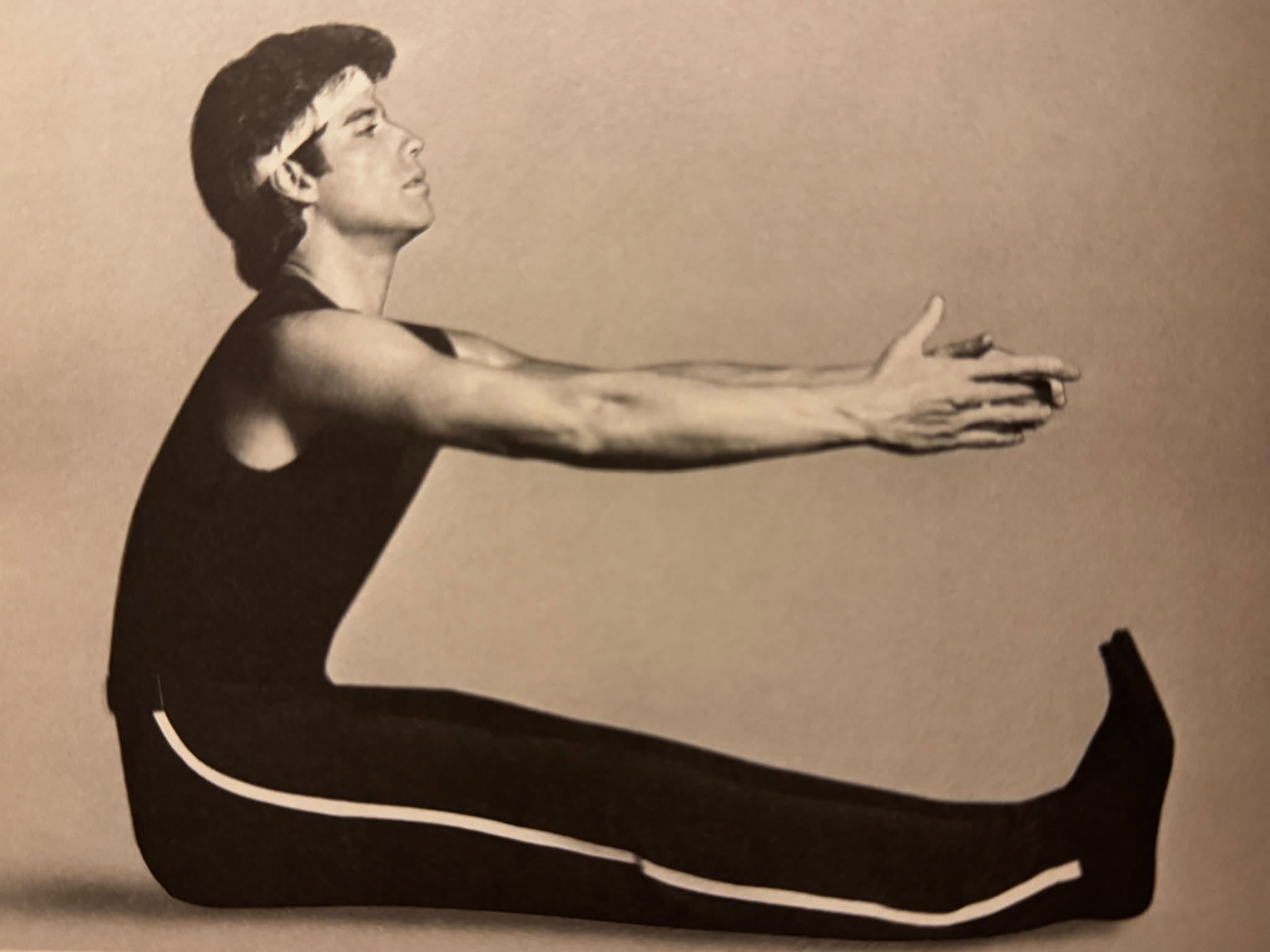 A black-and-white photo from a 1984 fitness book: a young John Travolta in a black sleeveless unitard and white headband sits on the floor with his legs extended straight in front of him, arms reaching forward over his legs mid-stretch.