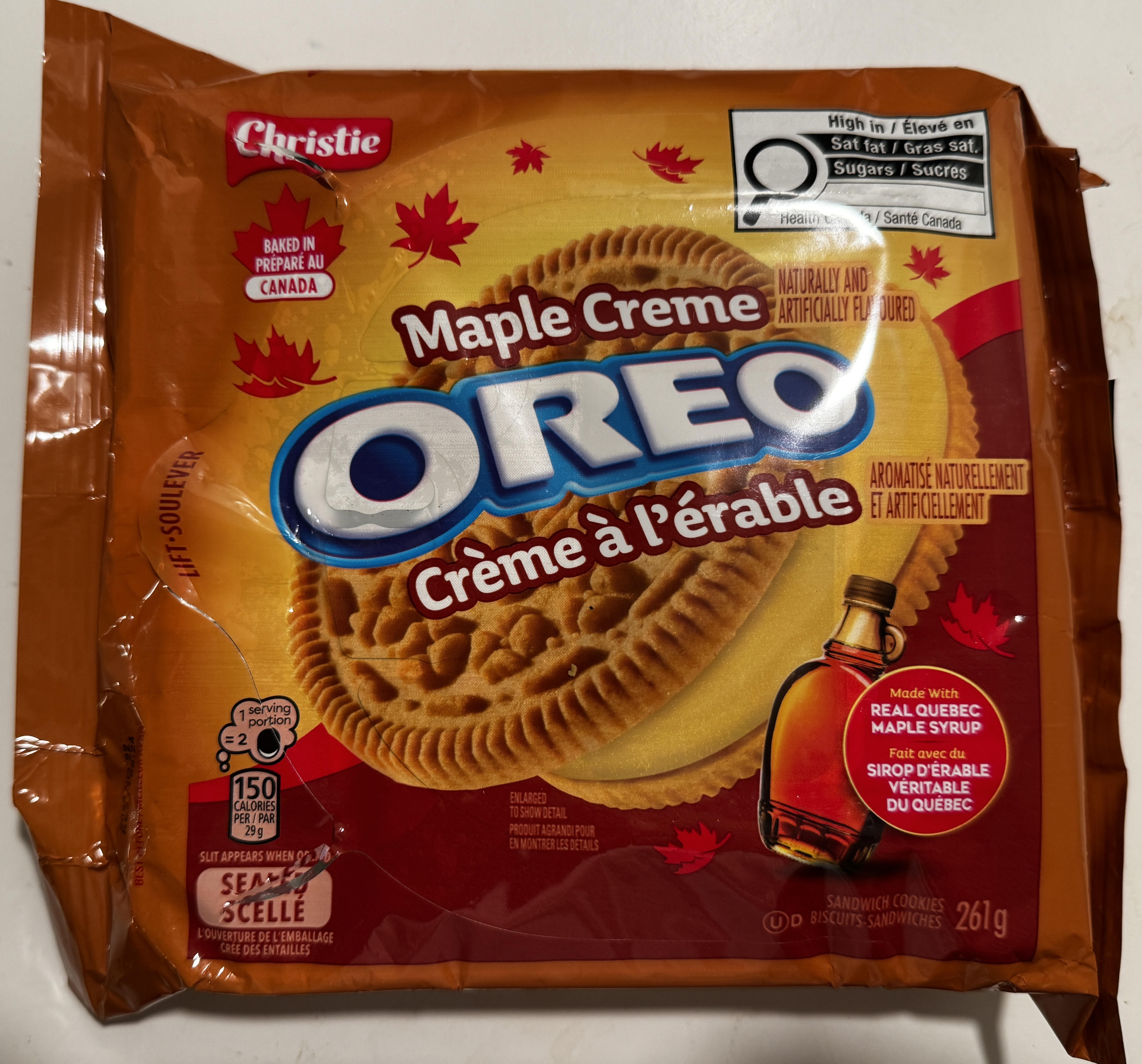 An orange Christie 'Maple Creme Oreo / Crème à l'érable' package baked in Canada, with golden sandwich cookies, scattered red maple leaves, a bottle of maple syrup, and a badge reading 'Made with Real Quebec Maple Syrup.'