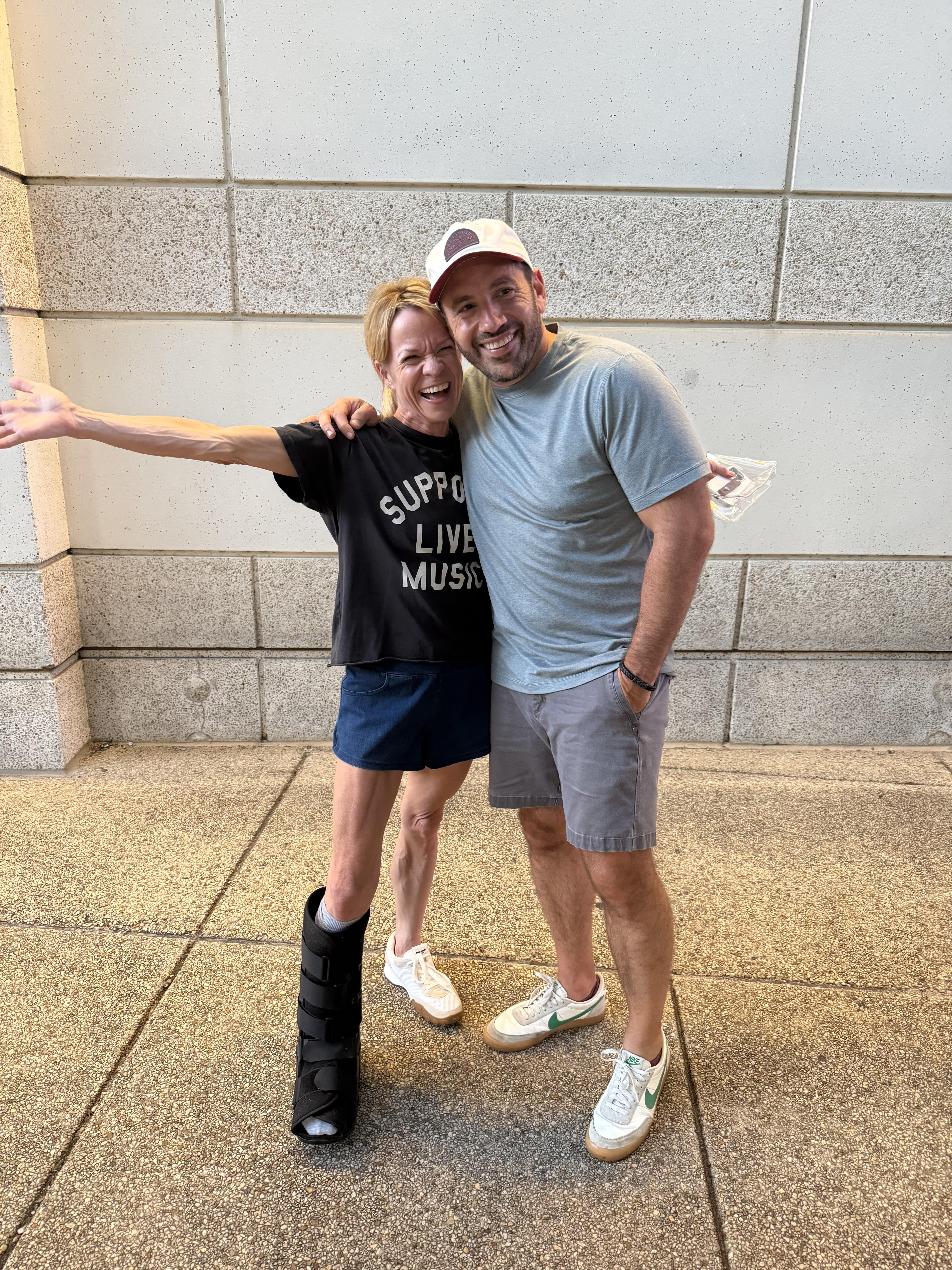 Me in a black 'Support Live Music' crop tee and blue shorts, boot on my right leg, one arm flung out and laughing, squeezed cheek-to-cheek with my orthopedic surgeon in a ball cap and gray tee outside the stadium.