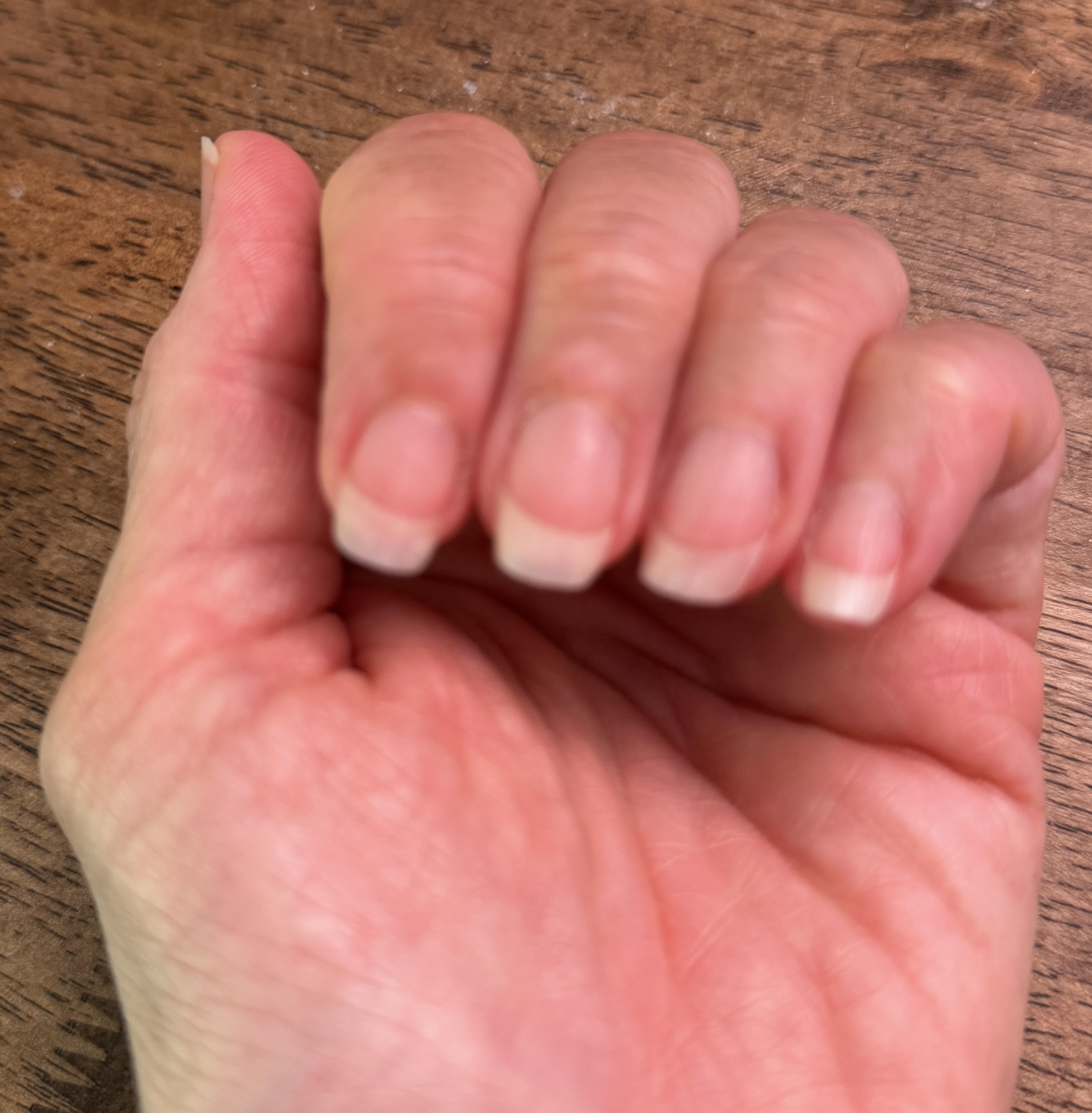 A close-up of the back of a woman's left hand resting on a wood table, fingers loosely curled, showing strong, smooth, healthy natural nails with pale-pink beds and clean white tips, freshly trimmed short.