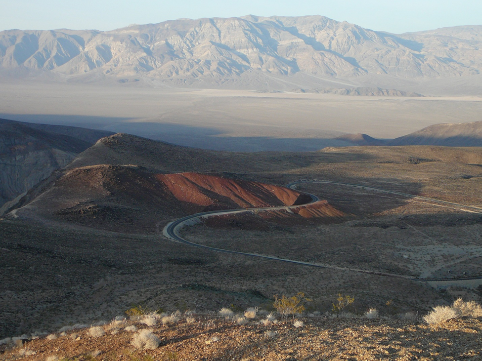 A narrow road winding down through dark, reddish volcanic hills into a vast open desert valley, with a long mountain range rising in the hazy distance; a remote Panamint Valley descent at low light.