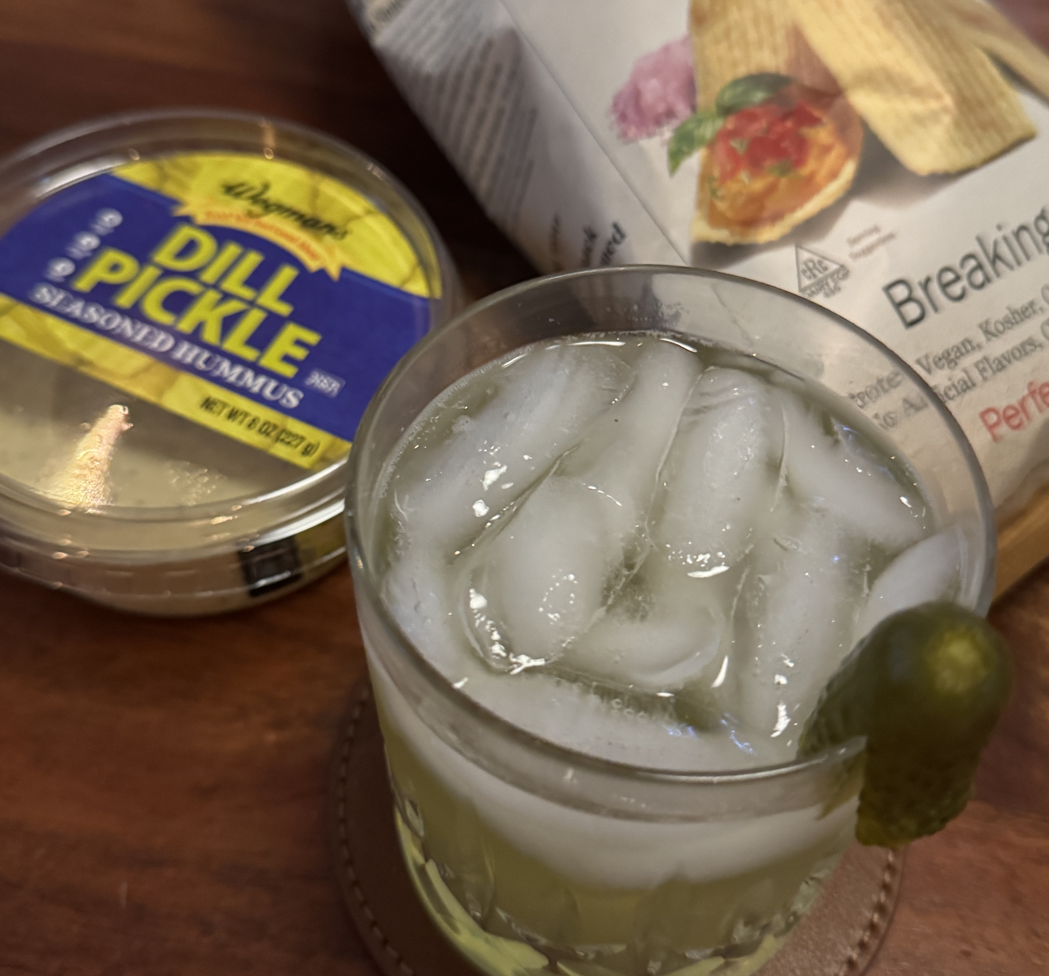 On a wooden table: a tub of Wegmans Dill Pickle Seasoned Hummus with a yellow and blue label, a rocks glass of pale green pickle mocktail over ice with a whole pickle perched on the rim, and a bag of crackers labeled 'Breaking Bread' resting behind them.