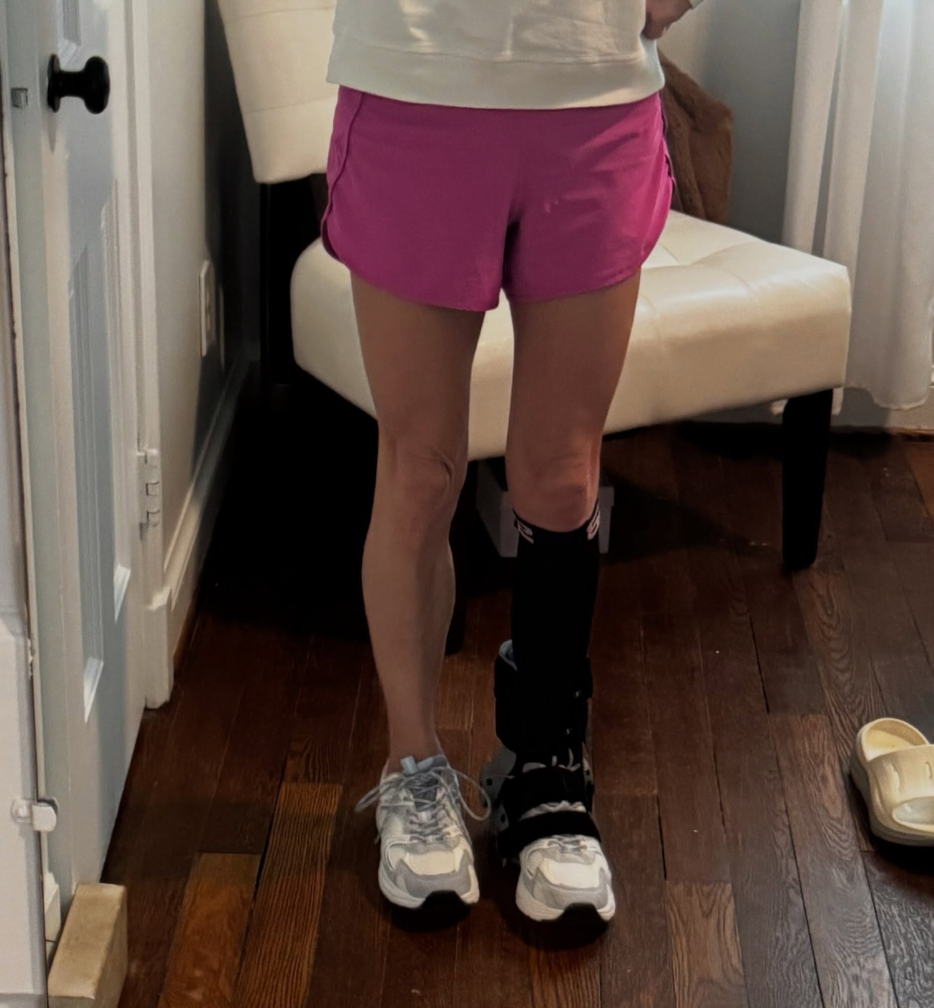 Standing in a hallway in pink shorts, both legs visible; the right leg wears a black knee-high sock and the low-profile RecoverX brace strapped over a white sneaker, next to a bare sneaker on the left foot.