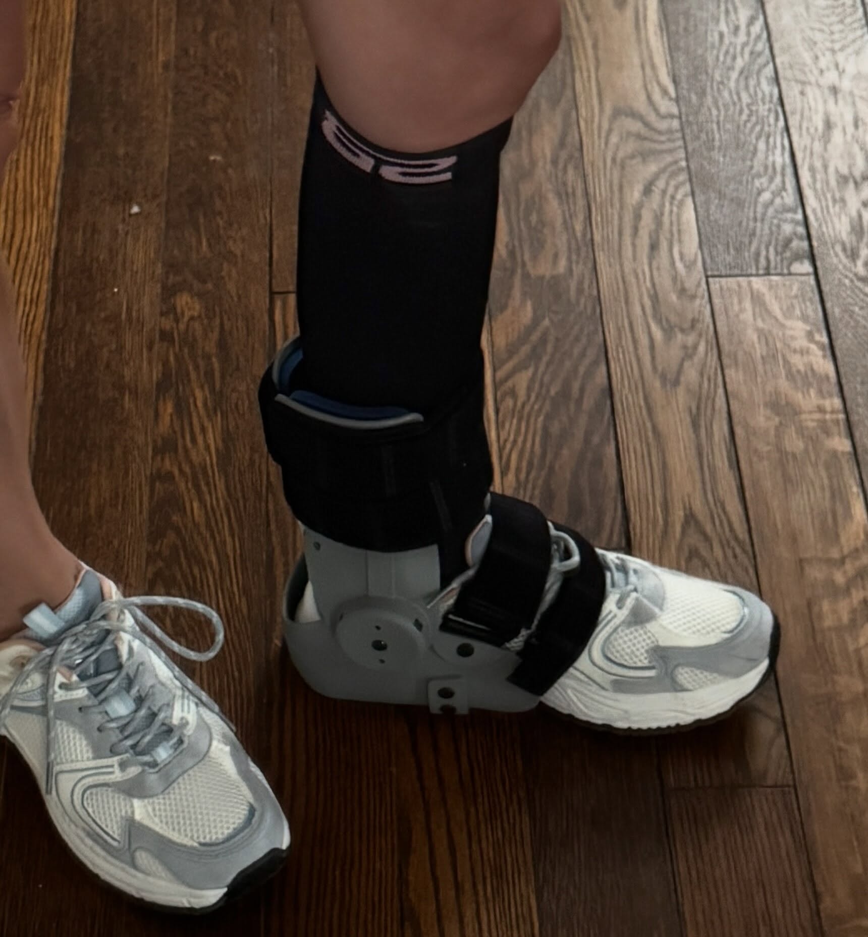 Close-up of the RecoverX brace on my right foot: a gray plastic footplate under the sole of a white sneaker, with a padded cuff and black straps cinched around the ankle and over the laces.