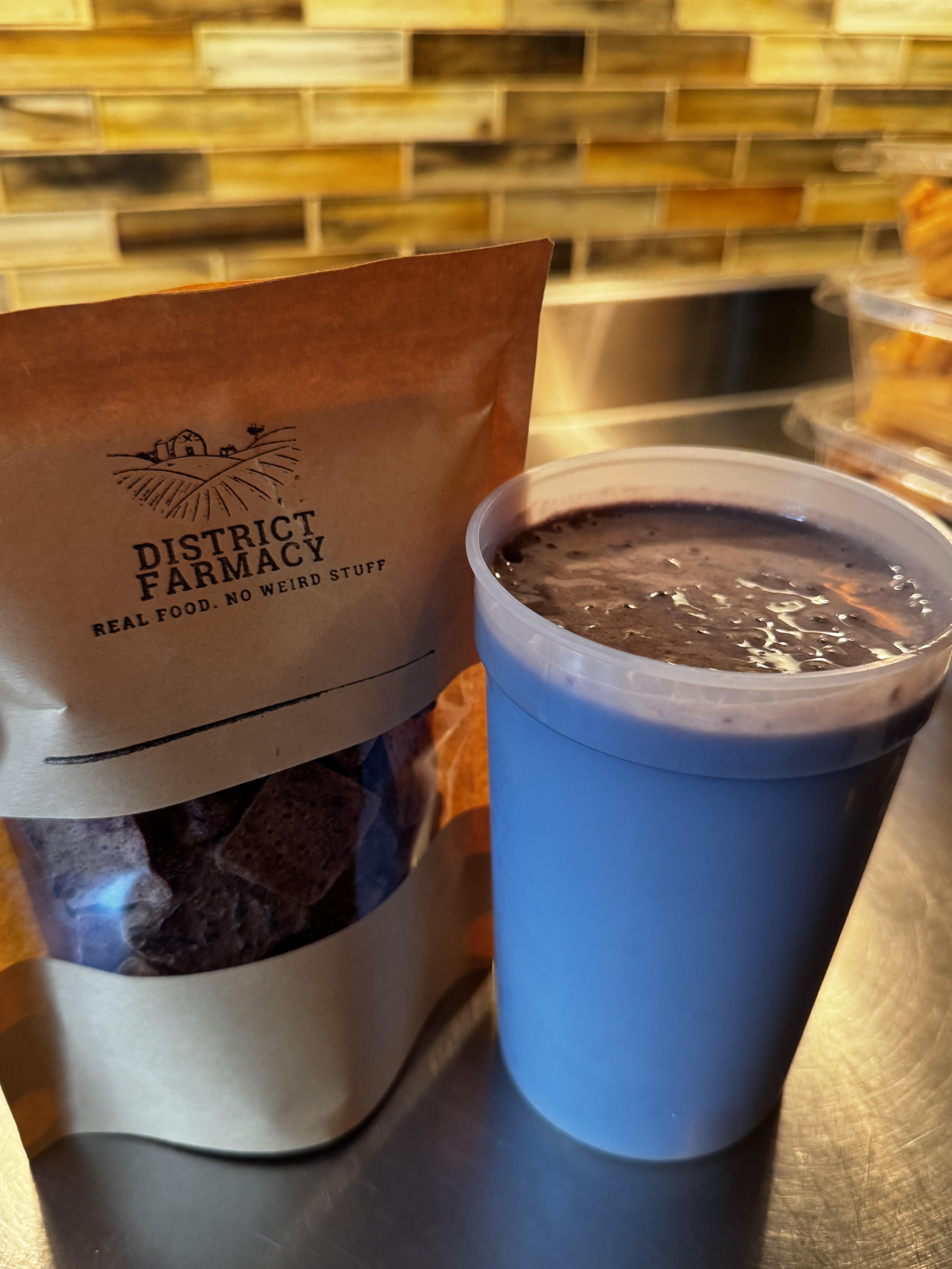 A resealable kraft-paper bag stamped 'District Farmacy — Real Food. No Weird Stuff' filled with dark purple freeze-dried blueberry squares, sitting on a stainless counter next to a tall blue cup of deep-purple protein shake, a gold-and-brown tile backsplash behind them.