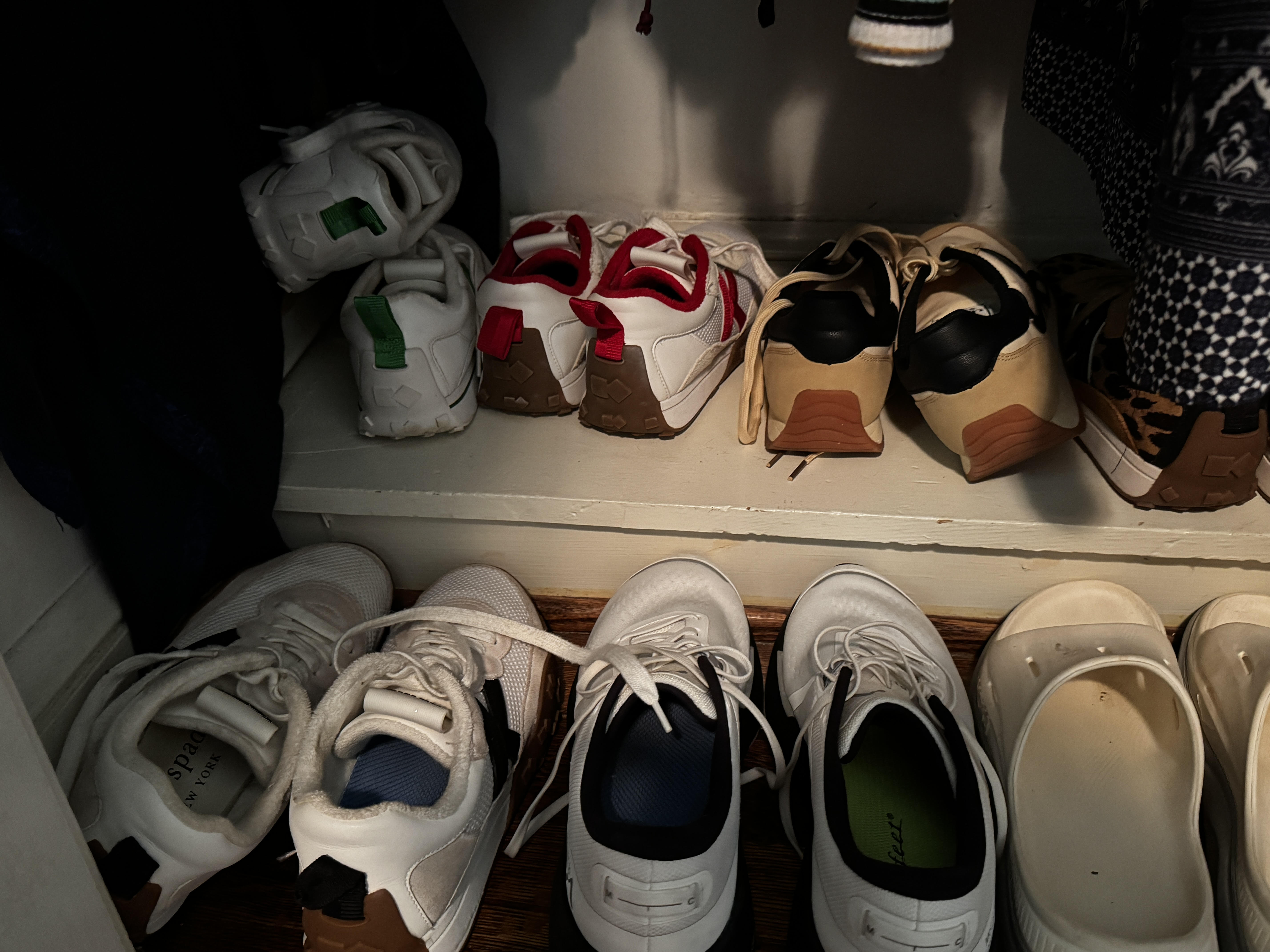 Two neat rows of athletic shoes lined up on a white closet shelf: white sneakers, a pair with red accents, tan suede trainers, and white slip-ons, with dark clothing hanging on either side.