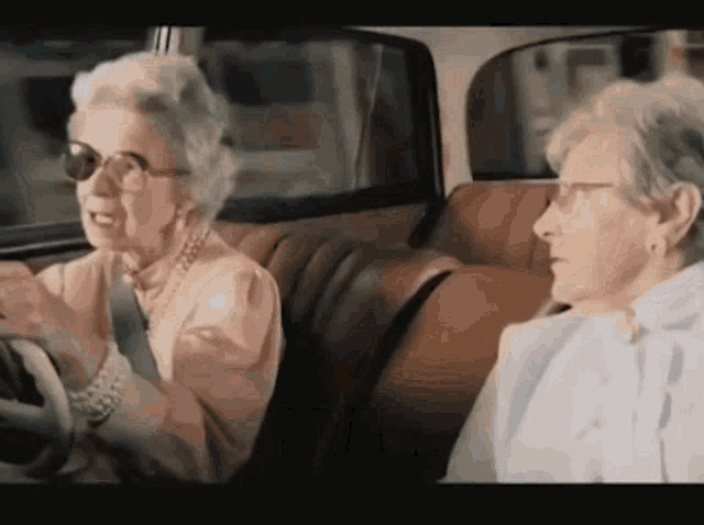 Two elderly women in the front seats of a car. On the left, a grey-haired woman in sunglasses and a pearl necklace grips the steering wheel and laughs; on the right, her friend in glasses and a white blouse turns to look at her. Still from the classic MiWay Insurance 'Muriel' ad.