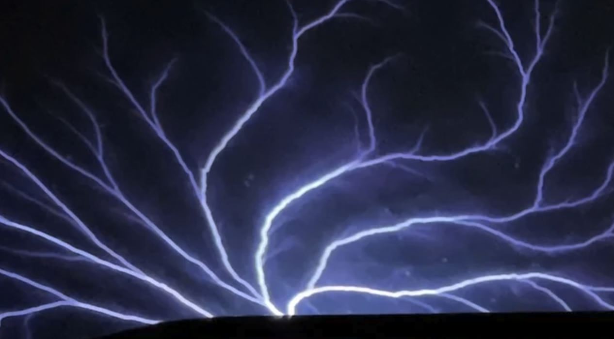 Bright purple-white electrical arcs branching upward from a dark horizontal surface against a black background, like a plasma discharge or St. Elmo's fire.