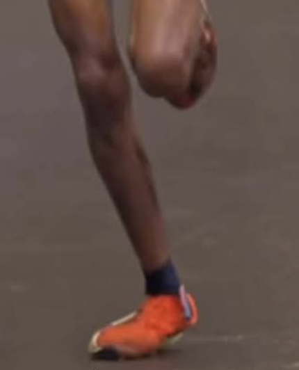 A close-up of a runner's lower leg mid-stride, the ankle collapsed dramatically inward so the orange shoe rolls onto its inner edge as the foot pushes off the pavement.