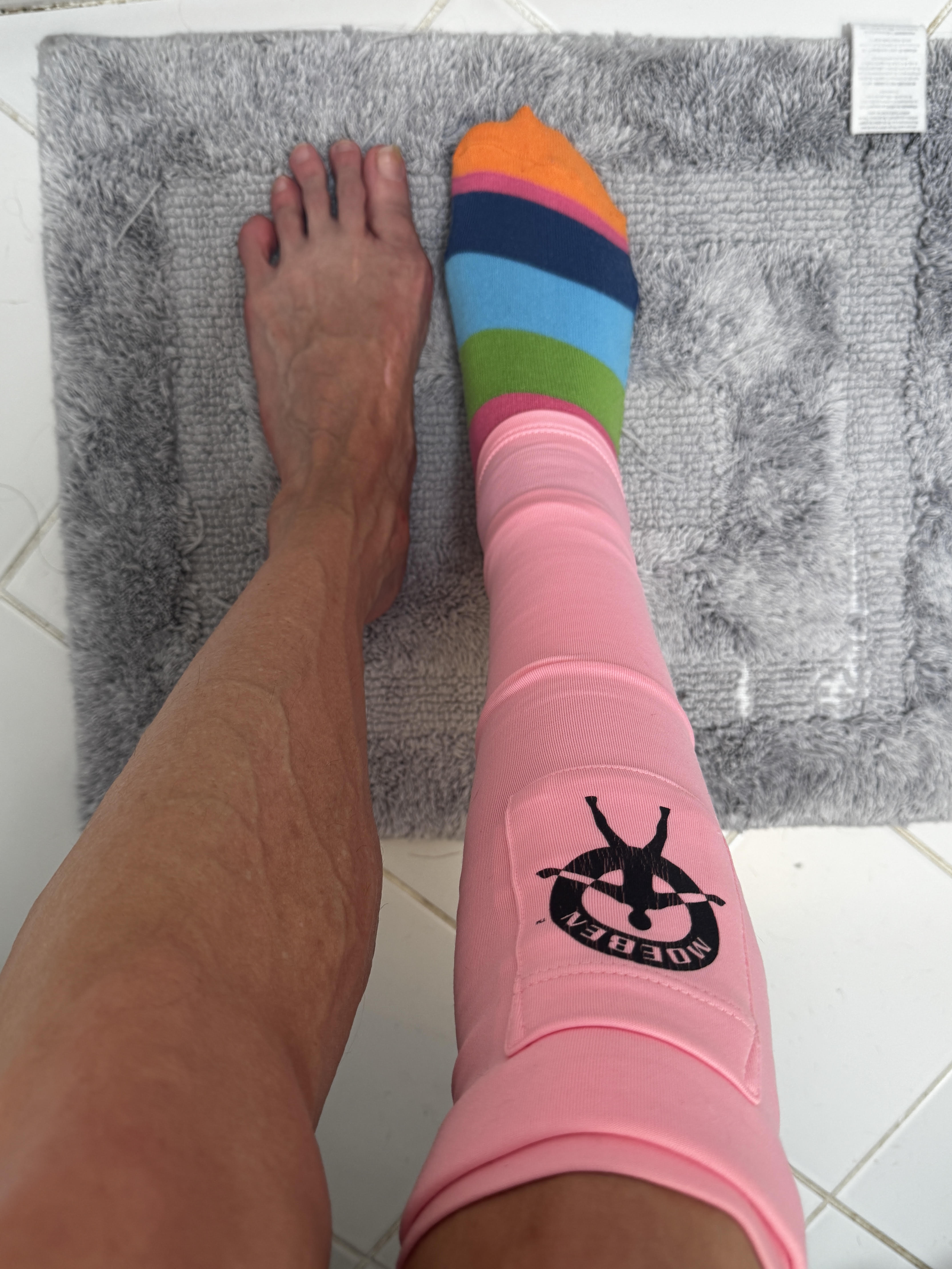 Top-down view of two bare legs on a gray bath mat over white tile. The right foot wears a bright rainbow-striped sock in green, blue, navy, pink, and orange, with a pink Moeben compression sleeve pulled up the shin, its black logo showing. The left foot is bare.
