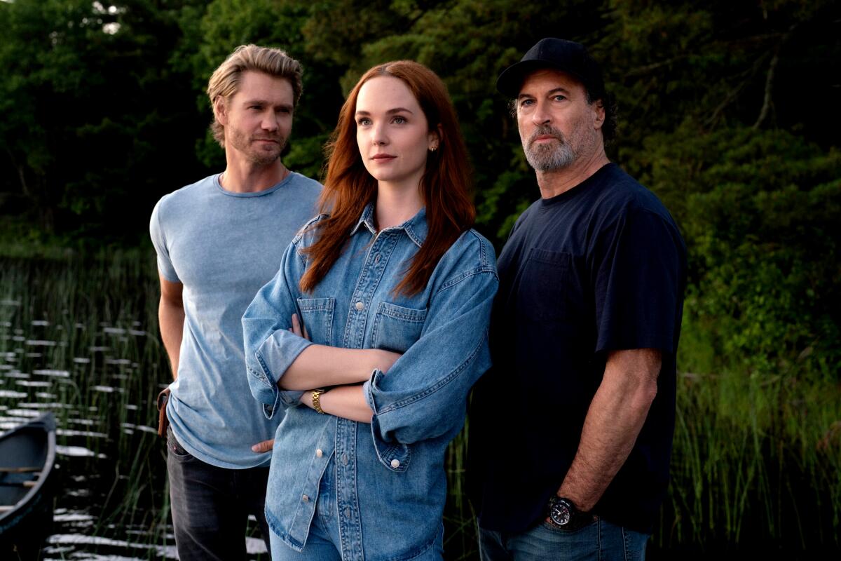Three cast members of Sullivan's Crossing stand at the edge of a lake at dusk: a blond man in a light blue t-shirt, a red-haired woman in a denim shirt with arms crossed, and a gray-bearded man in a dark t-shirt and black ball cap.
