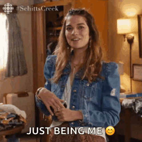 Alexis from Schitt's Creek in a denim jacket, smiling and shrugging, with the caption 'Just being me' and a smiley emoji.