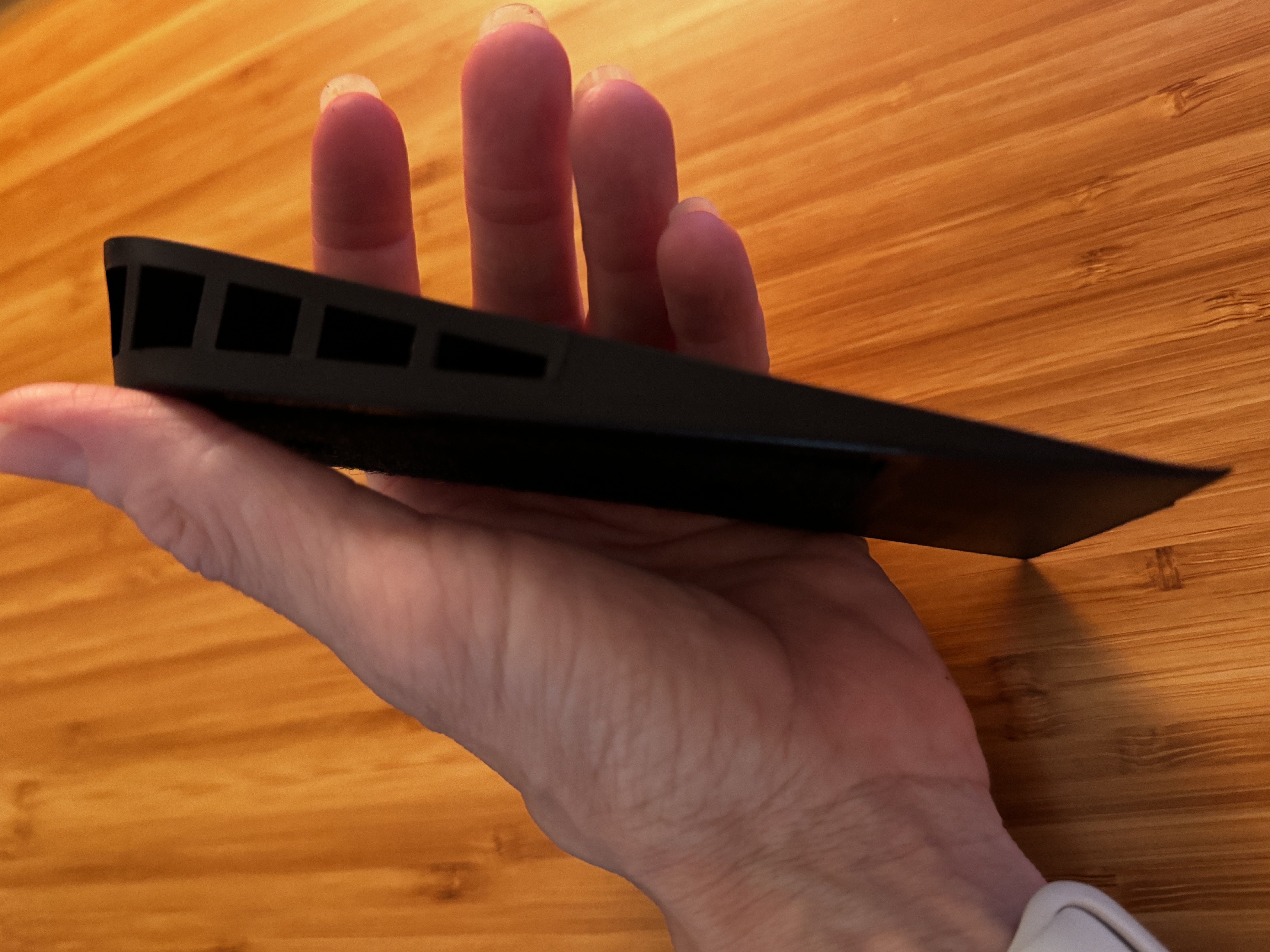 A hand holding a flat black boot wedge edge-on over a wood tabletop, its tapered heel-to-toe slope and hollow cutouts visible.