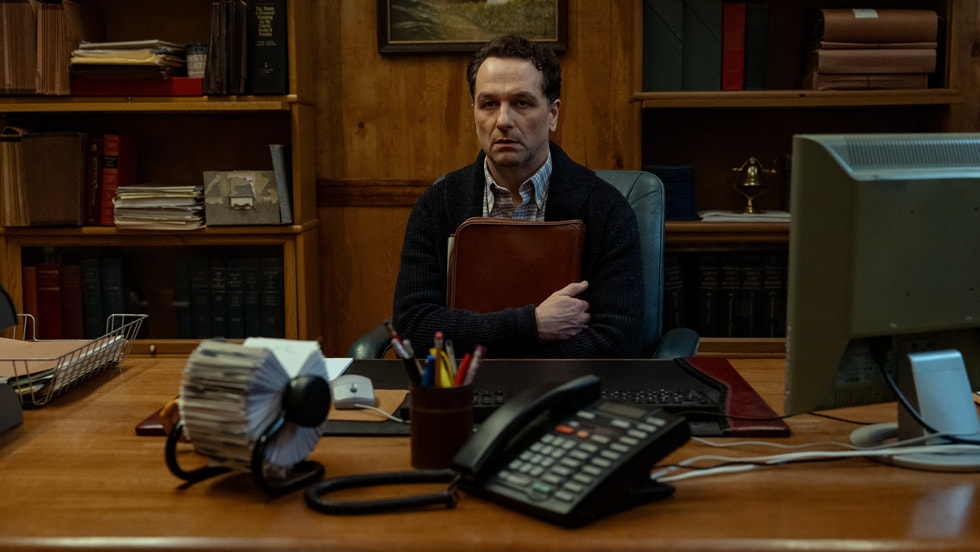 Matthew Rhys as a rumpled small-town official sits behind a cluttered wooden desk, hugging a leather folder to his chest with a wary expression, surrounded by bookshelves, a Rolodex, and an old office phone.