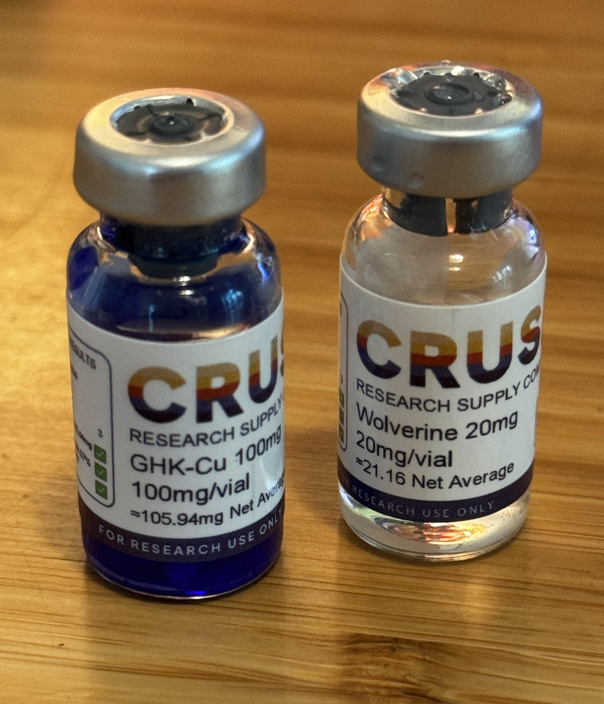 Two small glass research vials on a wood counter: a deep cobalt-blue vial labeled "GHK-Cu 100mg / 100mg/vial" on the left, and a clear vial labeled "Wolverine 20mg / 20mg/vial" on the right, both stamped "FOR RESEARCH USE ONLY."