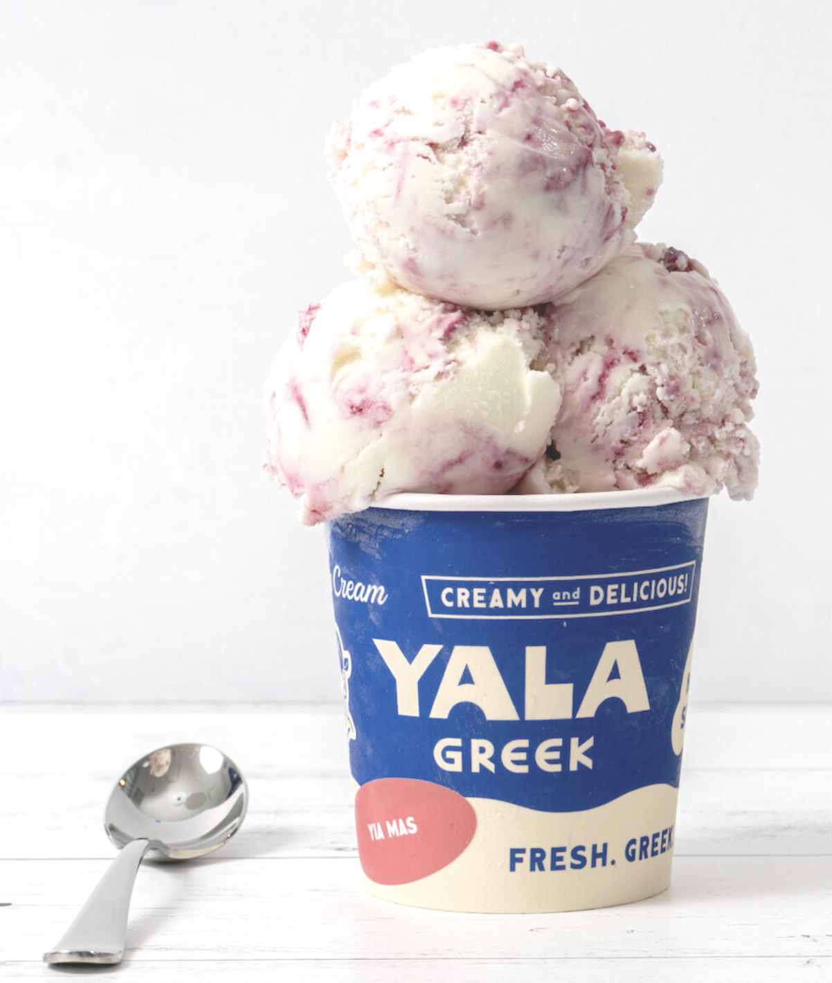 A pint of Yala Greek ice cream in a blue and cream container, overflowing with scoops of white ice cream swirled with berry ribbons, a silver spoon resting beside it.